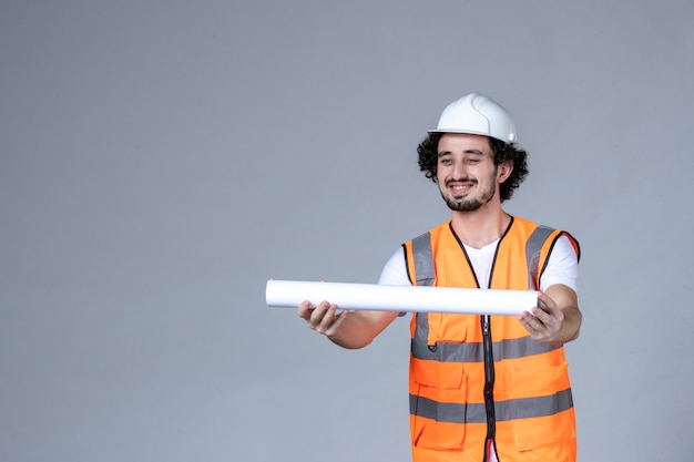 Front view of happy satisfied male constructor in warning vest wearing safety helmet and holding blank on gray wave wall