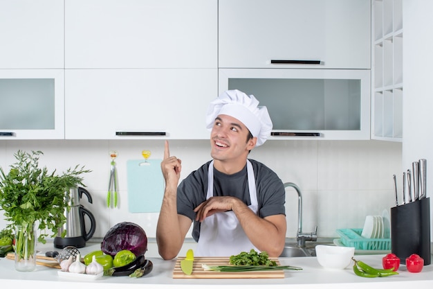 Front view happy young cook in uniform pointing at kitchen cupboard