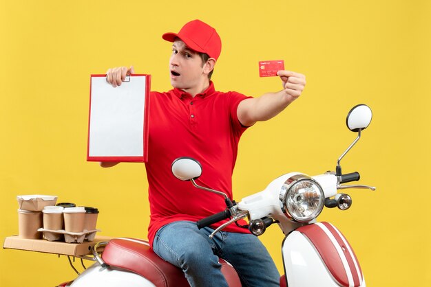 Front view of hardworking young guy wearing red blouse and bank card delivering orders holding document and bank card on yellow background