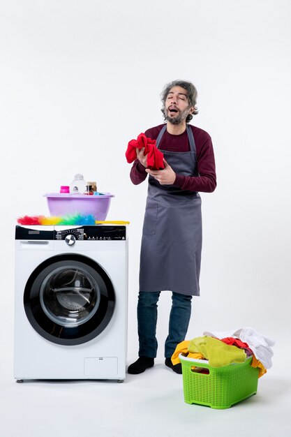 Front view housekeeper man with closed eyes holding red towel standing near washing machine on white background