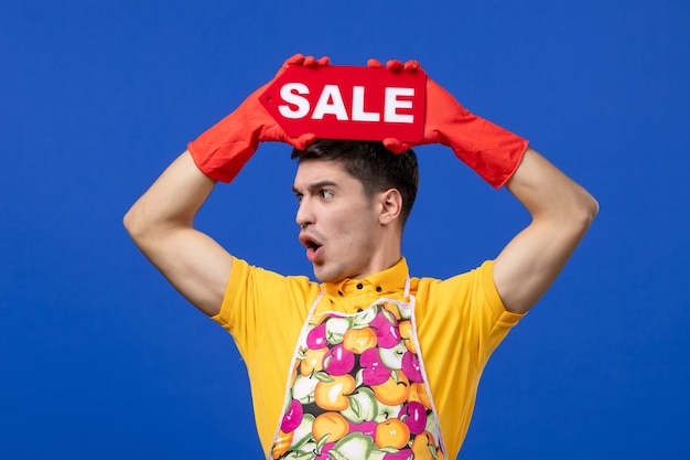 Front view housekeeper man in yellow t-shirt raising sale sign over head on blue space