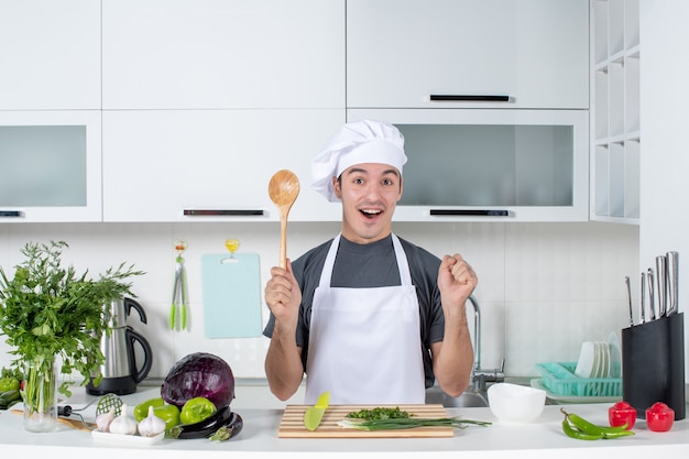 Front view joyful male chef in uniform holding wooden spoon behind kitchen table