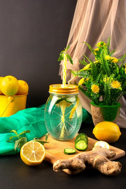 A front view lemon cocktail fresh cool drink inside glass cup sliced and whole lemons cucumbers along with flowers on the dark background cocktail drink fruit