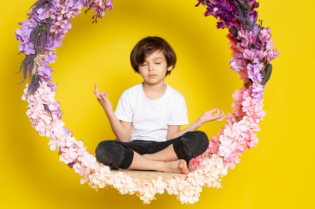 A front view little boy in white t-shirt sitting on the flower made stand on the yellow desk