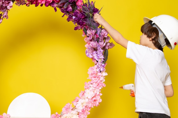 Free Photo a front view little boy in white t-shirt and white helmet decorating flower stand on the yellow desk