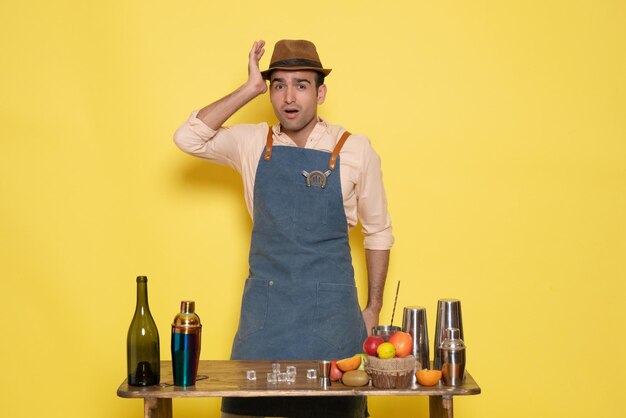 Front view male bartender in front of table with drinks and shakers on light yellow wall drink night alcohol bar color club