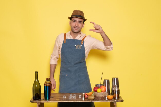 Front view male bartender in front of table with drinks and shakers on a yellow wall drink night alcohol bar color club male