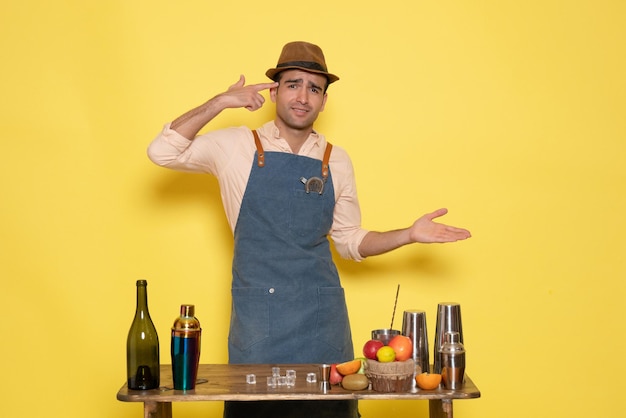 Front view male bartender in front of table with drinks and shakers on yellow wall drink night alcohol bar color club male