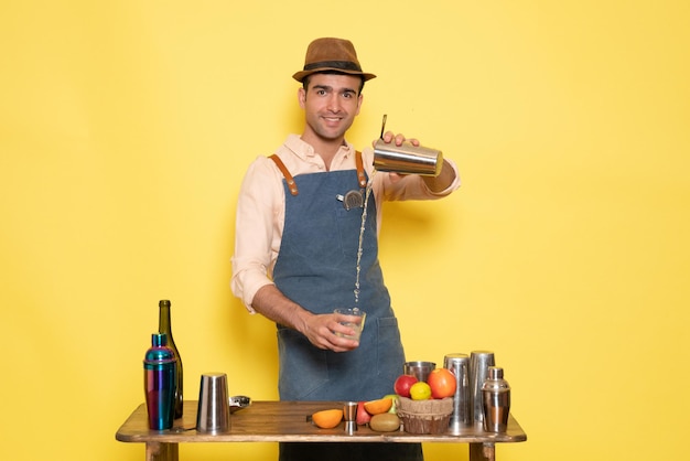 Front view male bartender in front of table with shakers and bottles making drink on yellow wall club bar drinks night alcohol