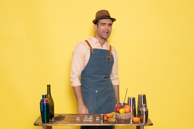 Front view male bartender standing in front of desk with drinks and fruits on the yellow wall night club bar male drink