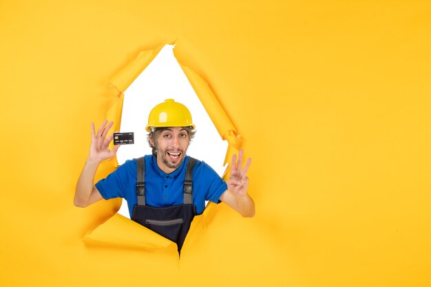 Front view male builder in uniform holding black bank card on a yellow background