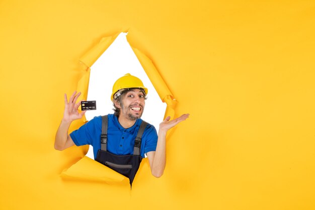 Front view male builder in uniform holding black bank card on yellow background