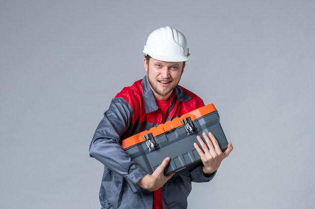 front view male builder in uniform holding heavy tool case on gray background