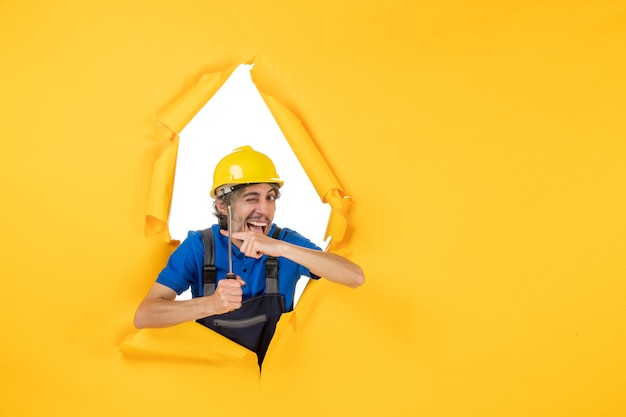 Front view male builder in uniform with screwdriver on yellow wall work constructor building color job worker architecture