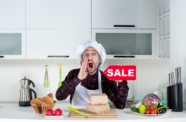 Front view of male chef in uniform holding up red sale sign saying something in modern kitchen