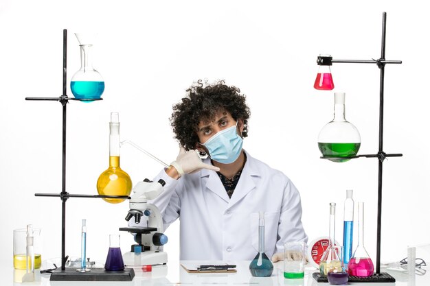 Front view male chemist in medical suit and with mask just sitting with different solutions posing on light white space