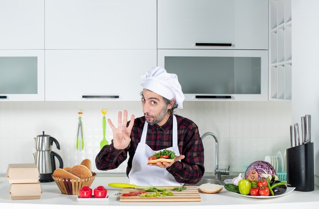 Front view of male cook holding up delicious burger standing behind kitchen table