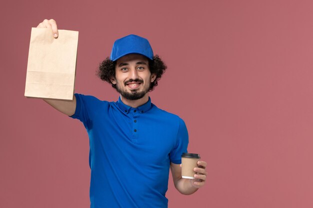 Front view of male courier in blue uniform and cap with delivery coffee cup and food package on his hands on pink wall