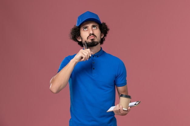 Front view of male courier in blue uniform and cap with delivery coffee cup and notepad on his hands writing notes on pink wall