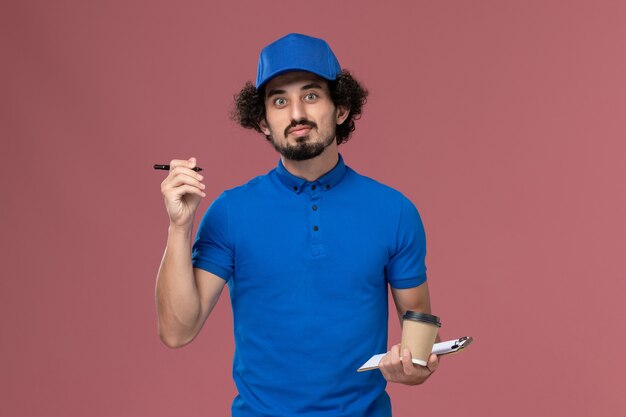 Front view of male courier in blue uniform and cap with delivery coffee cup and notepad on his hands writing notes on the pink wall