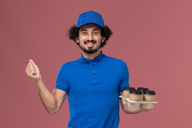 Front view of male courier in blue uniform cap with delivery coffee cups on his hands on light-pink wall