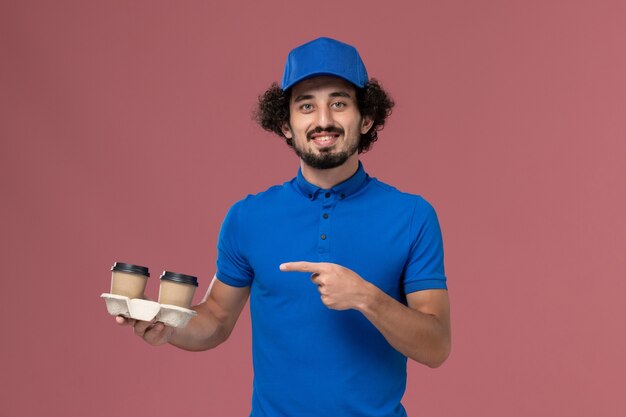 Front view of male courier in blue uniform and cap with delivery coffee cups on his hands on pink wall