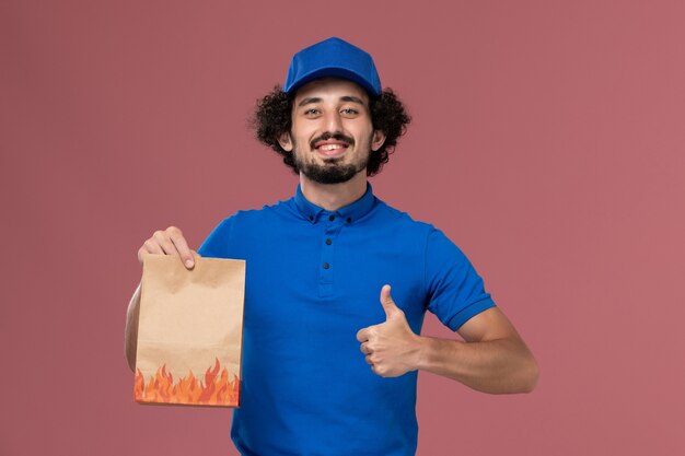 Front view of male courier in blue uniform cap with delivery paper food package on his hands on the light pink wall