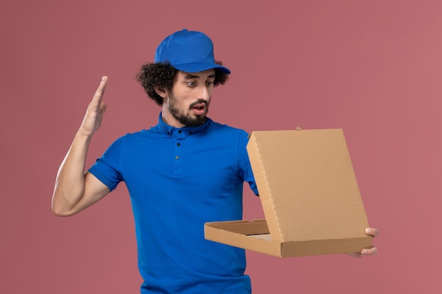 Front view of male courier in blue uniform cap with open food box on his hands on light-pink wall