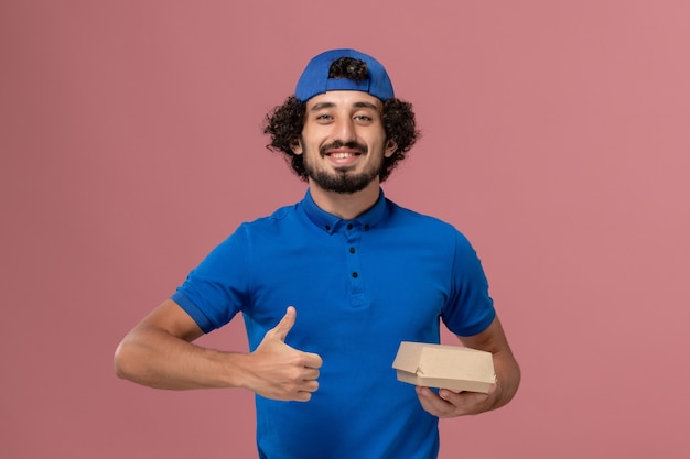 Free Photo front view male courier in blue uniform and cape holding little delivery food package on pink wall uniform delivery service company job employee