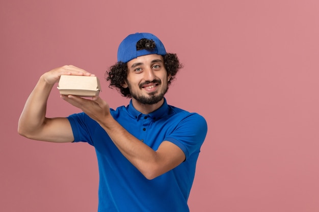 Free Photo front view male courier in blue uniform and cape holding little delivery food package on the pink wall