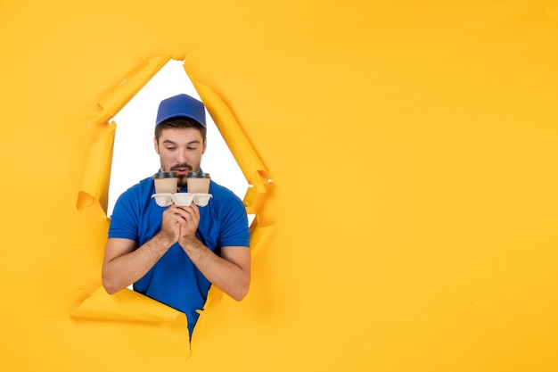 Free photo front view male courier in blue uniform holding coffee cups on the yellow space