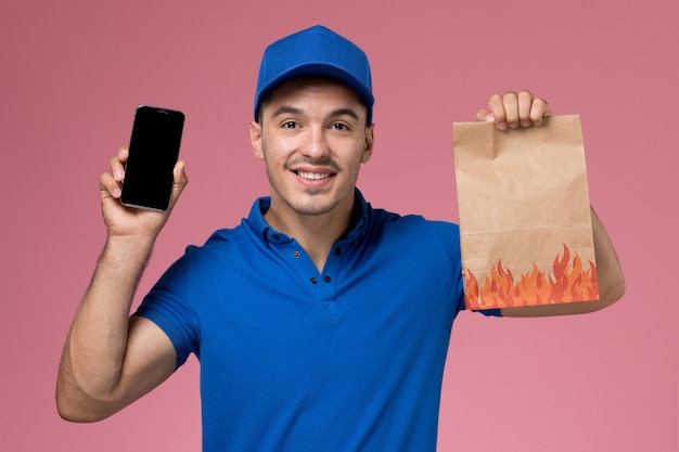 Front view male courier in blue uniform holding food package and phone on the pink wall, uniform service delivery