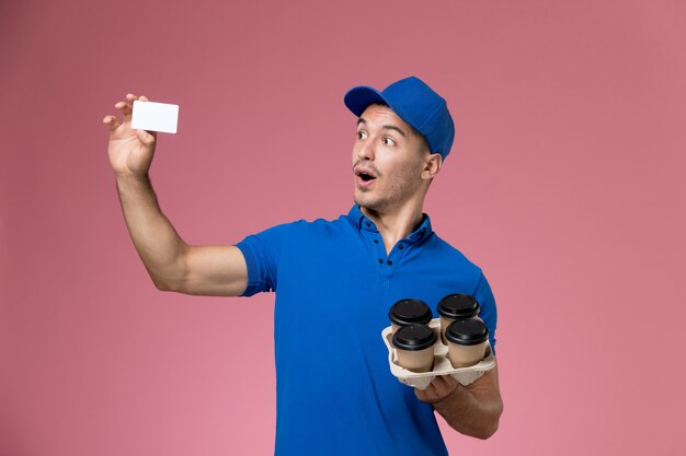 Front view male courier in blue uniform holding white card and delivery coffee cups on pink wall, uniform job worker service delivery