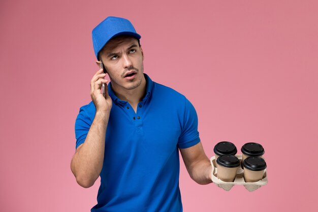 Front view male courier in blue uniform talking on the phone holding coffee on pink wall, worker uniform service delivery