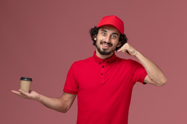 Free photo front view male courier delivery man in red shirt and cape holding brown coffee cup on pink wall service uniform delivery employee worker