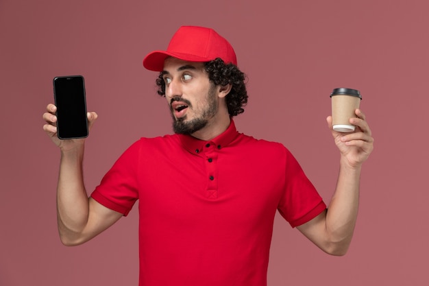 Front view male courier delivery man in red shirt and cape holding brown delivery coffee cup and phone on light pink wall service delivery employee job