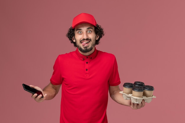 Free photo front view male courier delivery man in red shirt and cape holding brown delivery coffee cups along with phone on pink wall service delivery employee