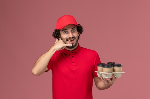 Front view male courier delivery man in red shirt and cape holding brown delivery coffee cups on the light pink wall male service delivery employee company