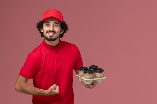 Free photo front view male courier delivery man in red shirt and cape holding brown delivery coffee cups on the light pink wall service work delivery employee