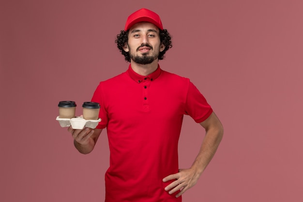 Free photo front view male courier delivery man in red shirt and cape holding brown delivery coffee cups on pink wall service delivery employee work