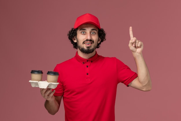 Front view male courier delivery man in red shirt and cape holding brown delivery coffee cups on pink wall service delivery employee worker