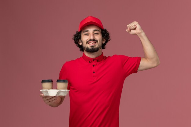 Front view male courier delivery man in red shirt and cape holding brown delivery coffee cups on pink wall worker service delivery employee