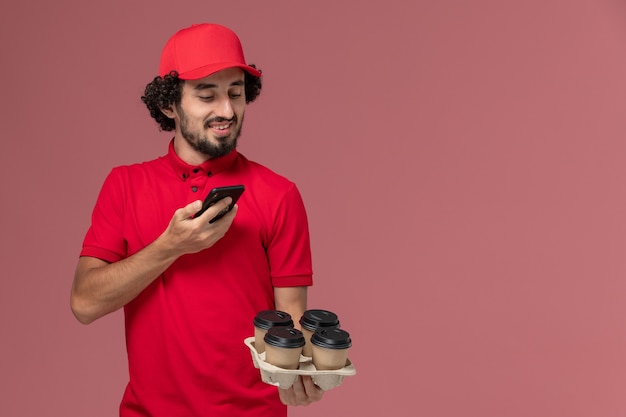 Free photo front view male courier delivery man in red shirt and cape holding brown delivery coffee cups taking photo on the light pink wall service delivery employee