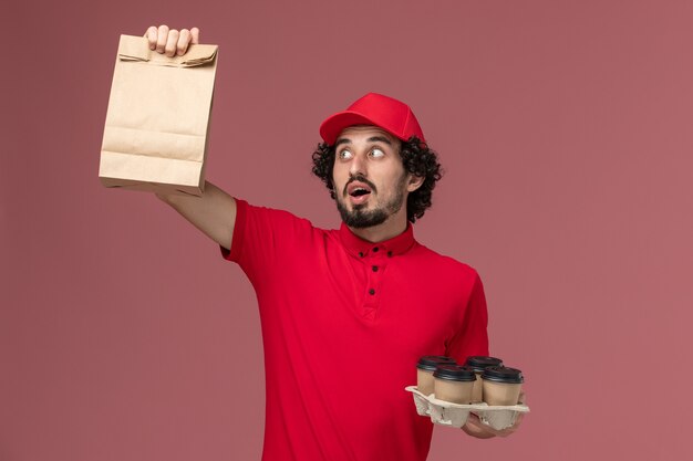 Front view male courier delivery man in red shirt and cape holding brown delivery coffee cups with food package on pink wall service delivery employee work