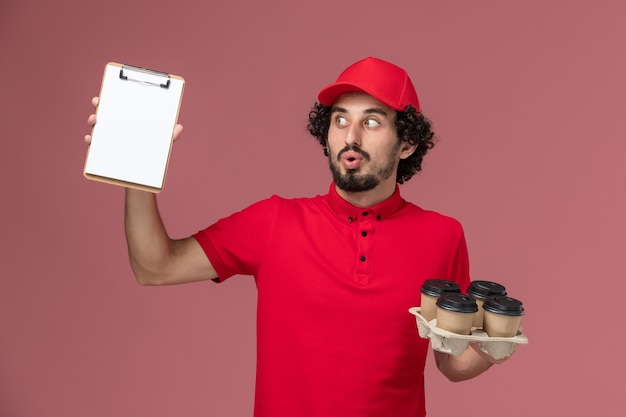 Front view male courier delivery man in red shirt and cape holding brown delivery coffee cups with little notepad on the pink wall service delivery employee male work