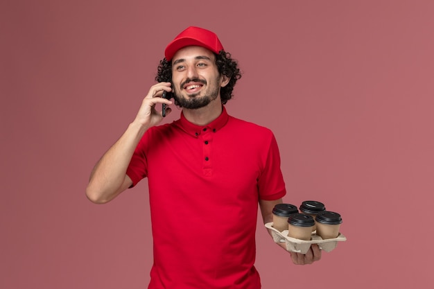 Free photo front view male courier delivery man in red shirt and cape holding coffee cups talking on the phone on the light pink wall male service delivery employee work