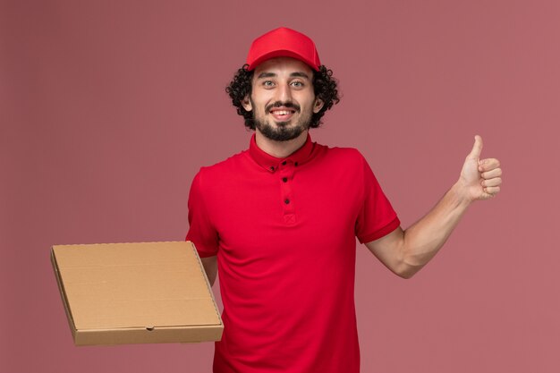 Front view male courier delivery man in red shirt and cape holding delivery food box on pink wall service delivery company employee work male