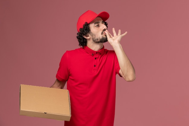 Free photo front view male courier delivery man in red shirt and cape holding delivery food box on pink wall service delivery company employee worker male