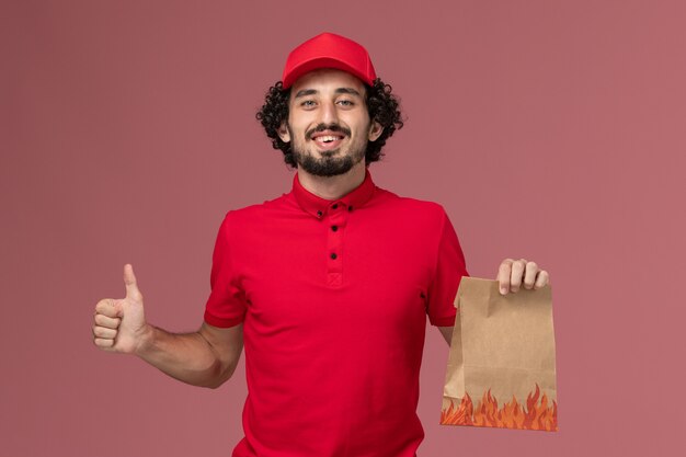 Front view male courier delivery man in red shirt and cape holding food package on pink wall service delivery company employee