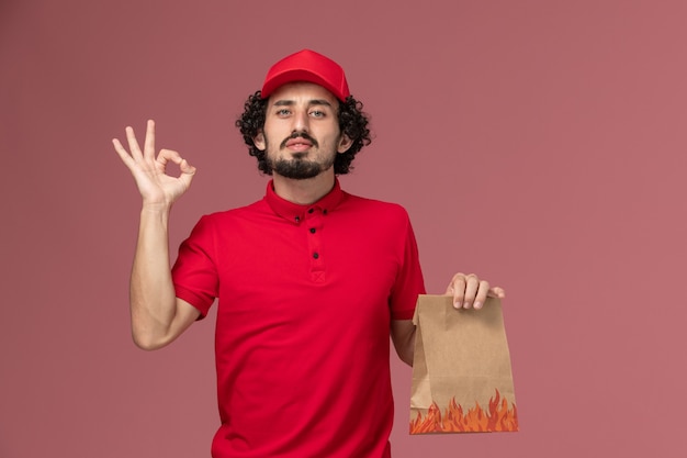 Front view male courier delivery man in red shirt and cape holding food package on pink wall worker service delivery company employee